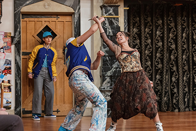 With their knives-holding hands upraised and crossed as Mercutio blocks Tybalt's thrust, Mercutio wearing a brown-patterend dress and bustier and white tennis shoes fights Tybalt in blue letter jacket with yellow trim and water-color style slacks. In the background, Sampson watcches by the stage door; he's also in a letter jacket and wearing a baseball cap, blue shirt, gray slacks, and blue tennis shoes. 