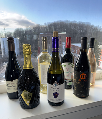 Accordia Wines-260228 Seven bottles of wine in two staggard rows on a window sill with a winter landscape through the window of buildings in front of a hill comprising bare trees. The wines are, from left: Red Earth Matchbook Petit Sirah, Astoria-Galie Prosecco, Domain Gavoty Grand Classique Vermentino, Vignamato "Violetta," Muse "Urania," and Baudry Dutour Chnino Rosé.