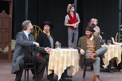 Lucio and the gentlemen sit at a saloon table as cowboys and saloon girs stand in the background along side wagon wheels and swining saloon doors.