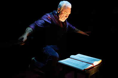 Patrick Page in dark purple shirt and black vest and pants, his face glowing in a spotlight, has his arms outstretched as he leans over an open book on the table.