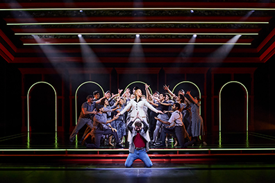 Eva Peron in a black trimmed white business jacket and dress stands with arms outstretched in a spotlight as the gray-clothed people of Argentina surround her in supplication while Che in red shirt and denim jacket and jeans knees with prayerful hands upraised at the front of the stage