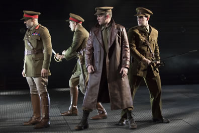 Othello and others stand in military uniforms with olive green service jackets, khaki jodhurs, brown boots, billed caps with red bands around them; two of the soldiers have their swords drawn; Iago stands in front, legs wide apart, in shin-length brown leather trench coat.