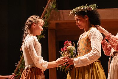 Eliza with braided pony tail hands Agnes standing across from her a boquet of just-picked flowers. Agnes in a nice Elizabethan dress is wearing Eliza's homemade crown of flowers on her head. Behind Agnes a pair of hands is adjusting her hair, and in the background is the wookd stuperstrugure decorated in fir garland